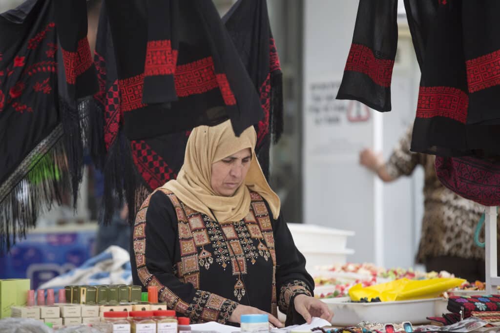 Femme au marché vêtue d'une robe à broderie traditionnelle et d'un hijab, entourée de châles et produits artisanaux.