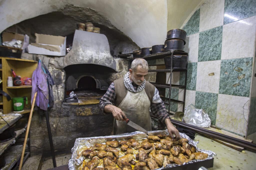 Cuisinier découpant du poulet rôti devant un grand four à bois traditionnel dans une cuisine en pierre.